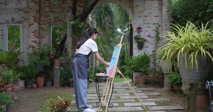 young asian woman artist wearing apron standing in garden painting calm scene on canvas during peaceful moment surrounded by nature expressing creativity through outdoor hobby