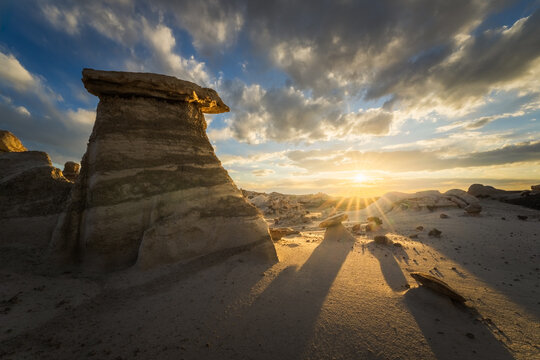 View of surreal rock formations casting long shadows in the desert landscape as the sun bursts on the horizon under a dramatic sky, Farmington, New Mexico, United States.