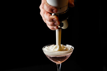 Close-up of a bartender pouring thick, white foam or whipped cream from a dispenser into a pink cocktail in a martini glass against a black background