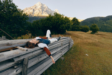 Obraz premium Woman lying on a wooden fence in a meadow with a mountain view, relaxing in nature among trees and open countryside, wearing casual jacket for a peaceful outdoor escape.