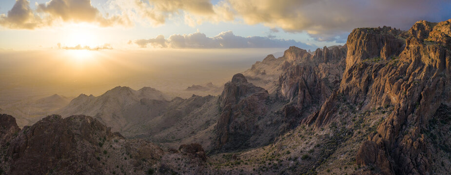 View of the sun's golden rays illuminating rugged, rocky peaks and casting long shadows across the arid landscape under a cloudy sky, Nipton, California, United States.