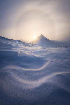 View of the sun rising behind a snow-covered mountain peak, with a halo effect visible in the sky above the undulating snowdrifts, Coldfoot, Alaska, United States.