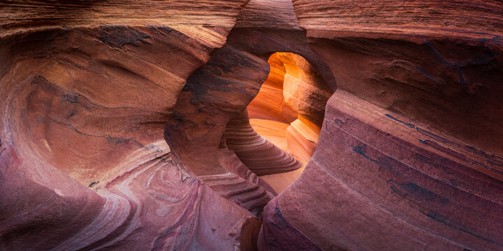 View of swirling, layered sandstone walls creating a natural tunnel, bathed in warm light filtering through the opening in Escalante, Utah, United States.