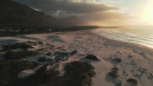 Aerial view of sandy beach meeting the ocean, with mountains looming in the background under a dramatic sky filled with clouds, Betty's Bay, Western Cape, South Africa.