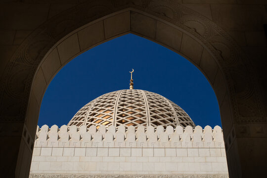 View of an ornate golden dome with a crescent moon atop a mosque, framed by an archway, Muscat, Oman.
