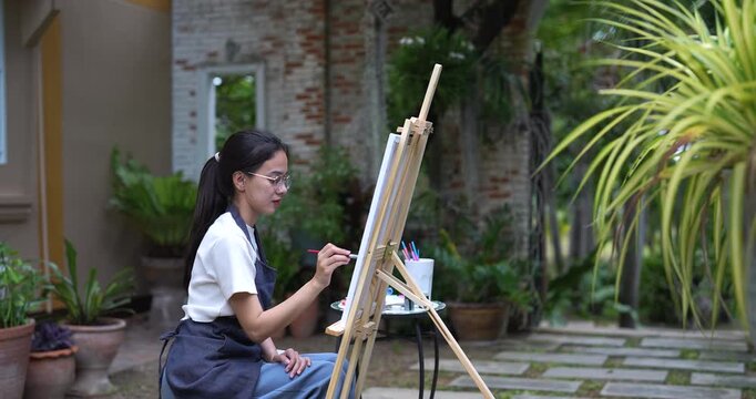 asian woman painting alone in garden sitting calmly in front of canvas focused on artwork enjoying tranquil moment of creativity surrounded by lush plants in peaceful backyard during quiet afternoon