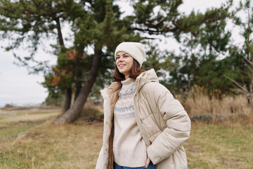 Obraz premium Woman in a warm beige coat and cream sweater stands in a windy park, wearing a knit beanie, casual winter fashion with trees and open field in the background.
