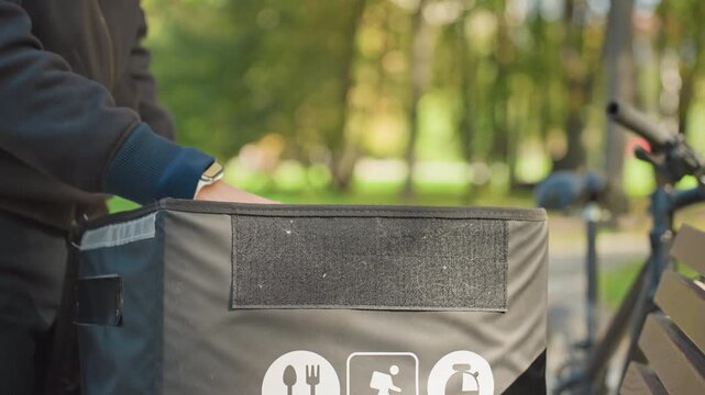 Bicycle courier packing food delivery bag, hands placing reusable container and foil wrapped meal into insulated bag at park pickup, sealing velcro flap, smartwatch visible, bicycle parked nearby,