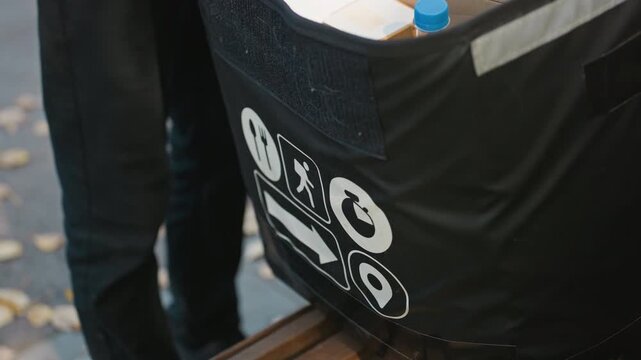 courier handling insulated delivery bag outdoors closeup, hand lifting flap to reveal food containers inside black thermal bag with white icons, reflective strip catching evening light, autumn leaves