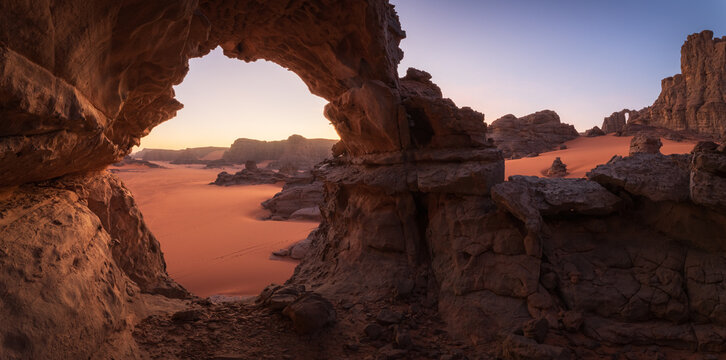 View of the sun kissing the red sands through a natural archway carved into the ancient rock formations, Djanet, Illizi Province, Algeria.