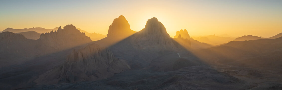 View of sunlit mountains casting long shadows across the rugged terrain, bathed in the golden light of sunrise or sunset, Djanet, Illizi Province, Algeria.
