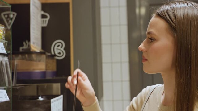 white woman sampling coffee at kiosk near counter in modern cafe selecting espresso from machine with touchscreen, thoughtful expression, tasting small sip, brunette hair, casual sweater, tiled wall