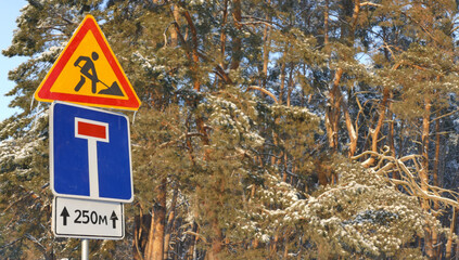 Signs indicating forest road repairs with distance indications for drivers against a backdrop of snow covered pine trees on a winter day
