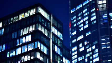 Obraz premium Big glowing windows in modern office building at night, in rows of windows light shines. Fragment of the glass facade of a modern corporate building at night. Modern glass office in city.