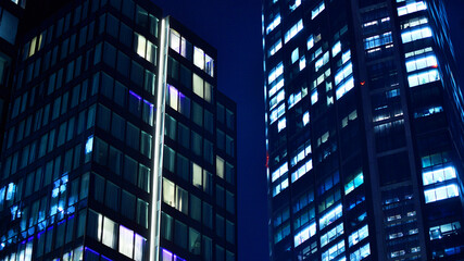Fototapeta premium Big glowing windows in modern office building at night, in rows of windows light shines. Fragment of the glass facade of a modern corporate building at night. Modern glass office in city.