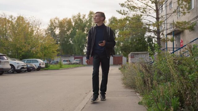 young man walking residential street checking phone. autumn suburban scene with parked cars, apartment building entrance, shrubs along sidewalk, casual jacket, focused expression, using delivery app,