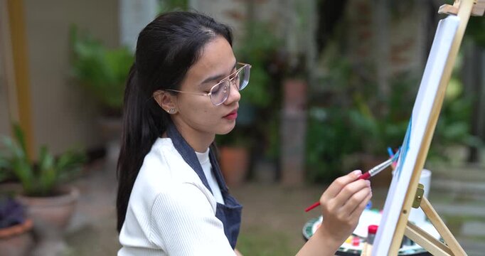 young asian woman holding brush painting on white canvas in backyard garden focusing on blue ocean scene expressing creativity through peaceful artistic moment in natural surrounding with concentratio