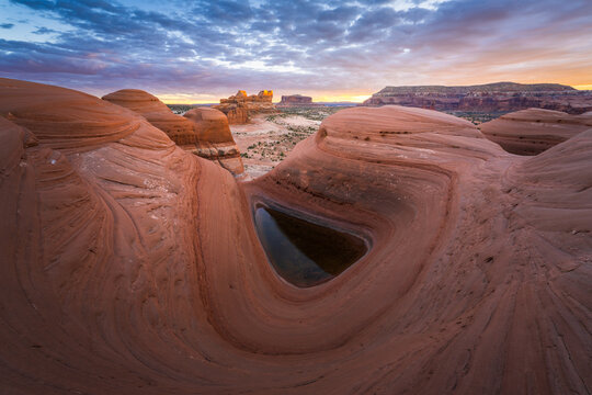 View of layered red rock formations embrace a still pool reflecting the sky's pastel hues at twilight in a desert landscape, Moab, Utah, United States.