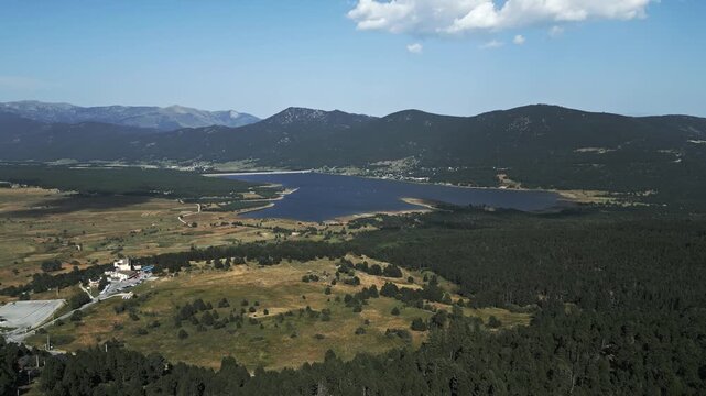 A sweeping drone shot capturing the vast blue waters of Lac de Matemale, surrounded by lush pine forests and the rolling peaks of the Capcir plateau in the French Pyrenees