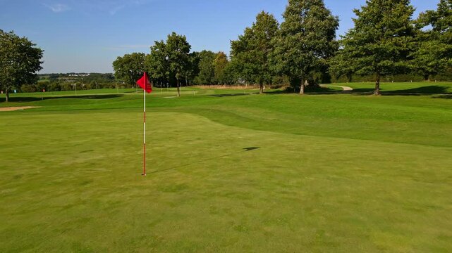 Golf green with flagstick and manicured grass under clear sky on quiet course, aerial pullback, empty nobody