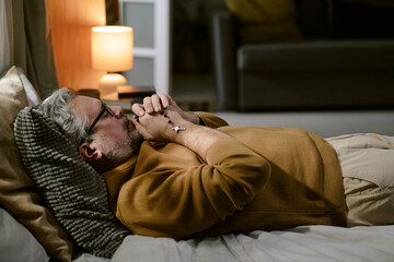 Obraz premium Middle aged Caucasian man lying on bed holding rosary beads with clasped hands near face appearing to pray or meditate, wearing glasses, relaxing in softly lit bedroom