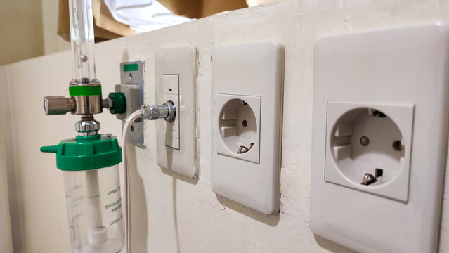 close-up shot of essential hospital wall equipment, including an oxygen flowmeter with humidifier bottle, medical gas or data port, and European-style power sockets (Type F)