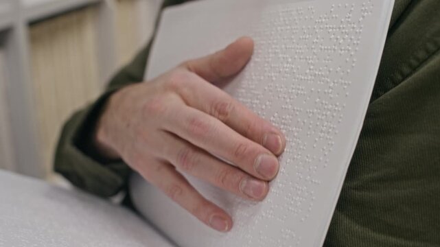 Cropped close up shot of hand of unrecognizable person with visual impairment standing in front of library shelf and reading braille book using fingers at high speed