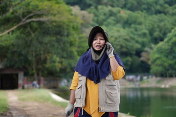 Naklejka premium Middle-aged Southeast Asian woman wearing a sun hat, yellow shirt, utility vest, and yellow boots takes a phone call while holding a clipboard, Gardening, landscaping, outdoor work, and agriculture