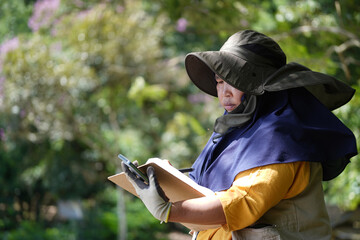 Obraz premium Middle-aged Southeast Asian woman in a sun hat, yellow shirt, utility vest, and yellow boots takes notes on her phone while surveying a forest. Gardening, landscaping, outdoor work, and agriculture
