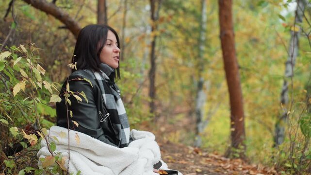 white woman sitting in autumn forest wrapped in blanket and wearing leather jacket and scarf, gazing into distance with reflective expression, leaves falling around, birch and pine trees