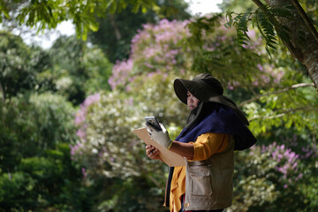 Middle-aged Southeast Asian woman in a sun hat, yellow shirt, utility vest, and yellow boots takes notes on her phone while surveying a forest. Gardening, landscaping, outdoor work, and agriculture