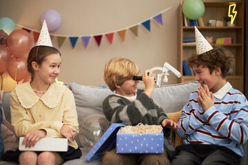 Fototapeta premium Three Caucasian children sitting on sofa celebrating birthday, boy in center using toy robot while girl and boy wearing party hats watching and smiling, balloons and decorations in background