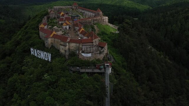 Aerial view of the Rasnov Citadel, perched atop a hill surrounded by dense green forests, with a funicular visible, Rasnov, Judetul Brasov, Romania.