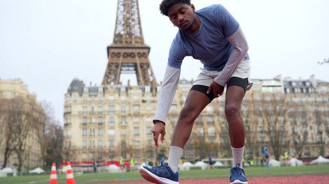 Young athletic man performing hamstring stretches on a running track, preparing for his training workout with the eiffel tower visible in the background on an overcast day in paris, france