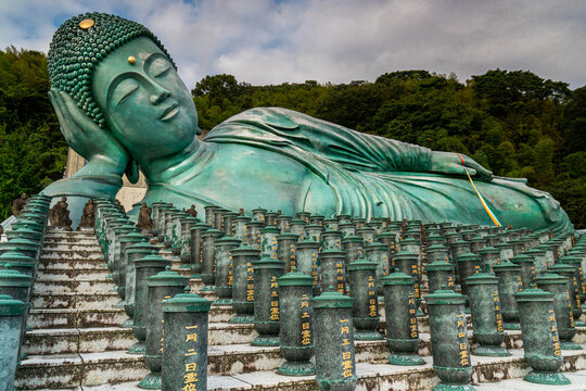 View of a monumental bronze Reclining Buddha resting serenely atop rows of stone pillars against a backdrop of lush forest, Nanzoin Temple, Sasaguri, Fukuoka Prefecture, Japan.