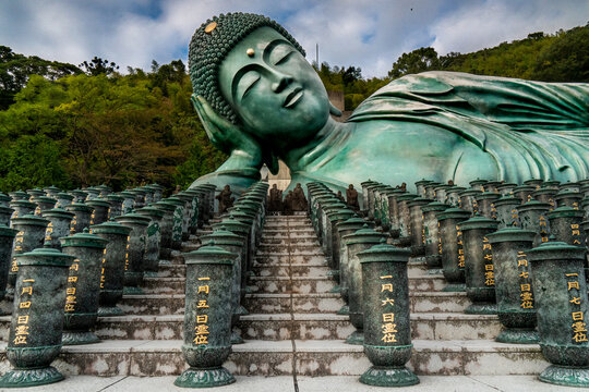 View of a colossal bronze Reclining Buddha statue rests serenely above stone steps adorned with ornate pillars in Nanzoin Temple, Sasaguri, Fukuoka Prefecture, Japan.