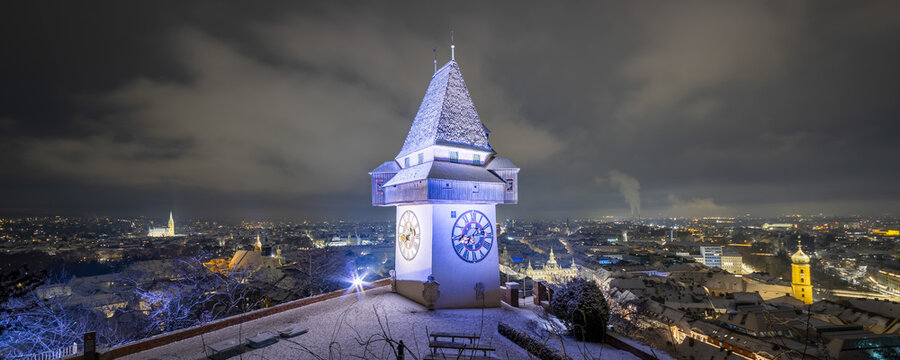 View of the iconic clock tower illuminated against the night sky, standing tall over the cityscape dusted with snow, a picturesque scene, Graz, Styria, Austria.