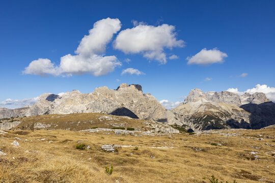View of rugged, sun-drenched mountains pierce the azure sky, casting long shadows across the golden, grassy foreground, Monte Piana, Trentino-South Tyrol, Italy.
