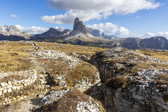 View of rugged stone trenches cutting through fields of golden grass, leading towards towering peaks under a vast sky, Monte Piana, Trentino-South Tyrol, Italy.
