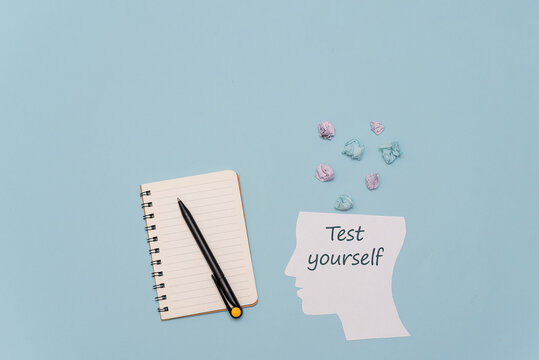A flat lay of a blank notebook, a black pen, and a paper cutout of a head with the text 'Test yourself'. Colorful paper stars are scattered around on a blue background.