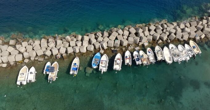 High-angle aerial view of several small motorboats moored alongside a concrete block breakwater in clear turquoise sea water.