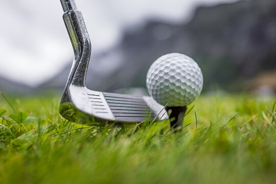 Golf club and ball on wet green field with mountains in background.
