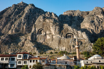 Ancient rock-cut tombs carved in cliff face above Amasya city with traditional Ottoman houses, mosque with minaret below in valley, heritage and historical architecture concept