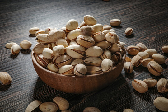 Wooden bowl filled with pistachios on dark wooden table, tasty salted nuts scattered around, healthy snack and natural food concept.