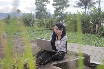Young Asian Muslim woman wearing a hijab talks on a smartphone while working outdoors with a laptop. Remote work, digital nomad, and work-from-anywhere lifestyle concept.