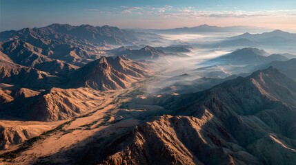 Aerial drone view captures a rugged desert landscape with rocky mountains, sand dunes, and soft morning mist in the vast valleys.