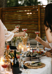 Friends raising coupe glasses with red cocktails around a candlelit outdoor table, enjoying an intimate dinner celebration with shared food and warm evening atmosphere.