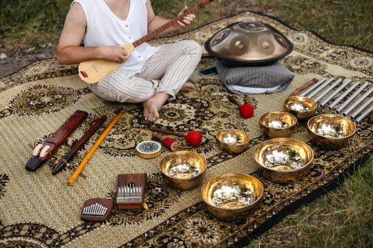 Wide view of a musician seated outdoors surrounded by sound healing instruments, including handpan, singing bowls and flutes, creating a serene wellness atmosphere.