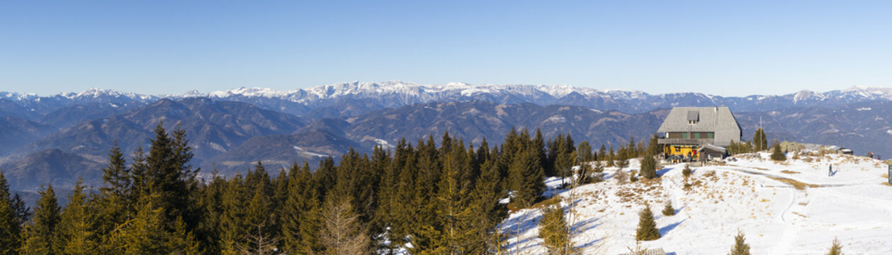 View of a cozy wooden mountain cabin nestled amidst snow-dusted slopes and towering pine trees, with snow-capped mountains in the distance, Bruck an der Mur, Austria.