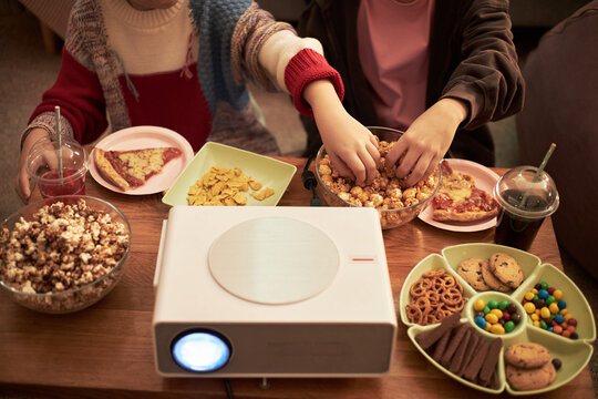 Two children reaching for popcorn and snacks while sitting together in front of projector, sharing pizza, cookies, pretzels, candy and drinks during movie night at home